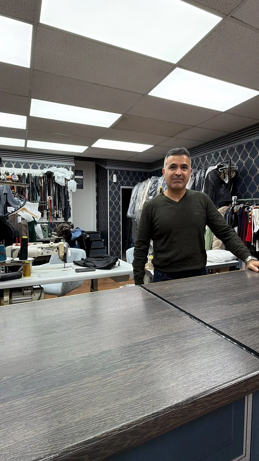 The tailor standing behind the counter at Best Stitch, with thread reels on the wall behind him